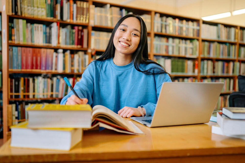 Smiling woman at library table books pen laptop